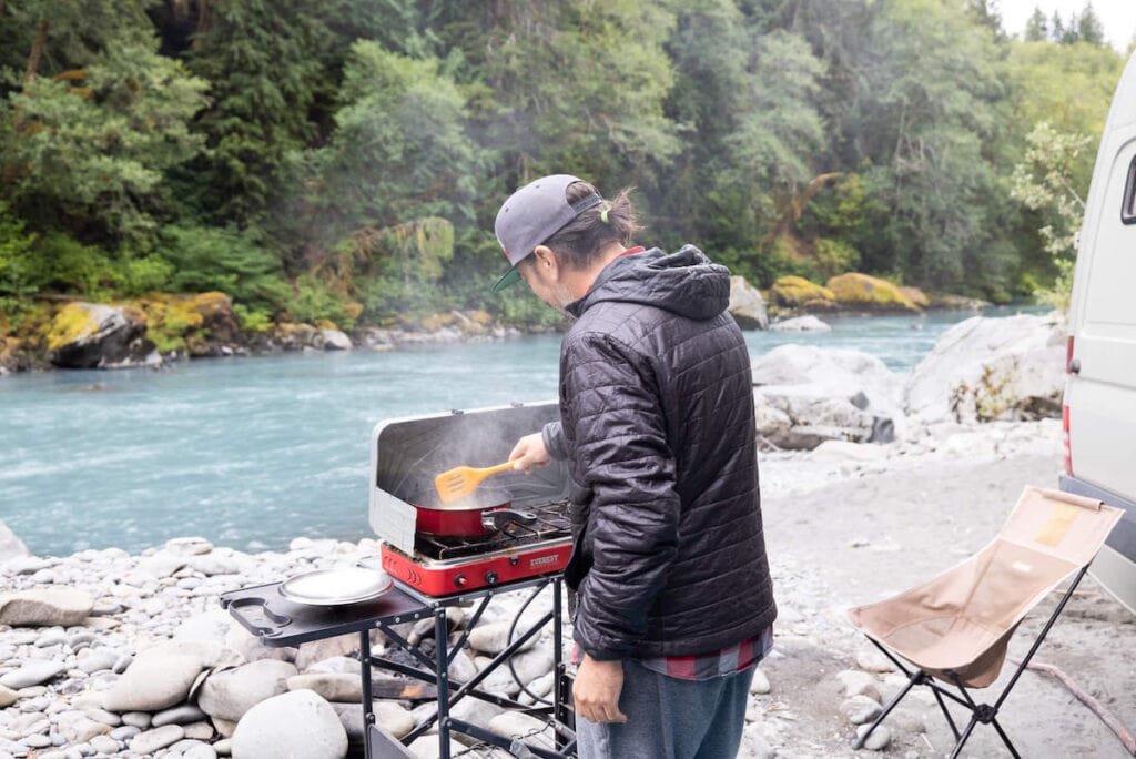 Чоловік готуємо їжу на вуличній плиті перед річкою в кемпінгу