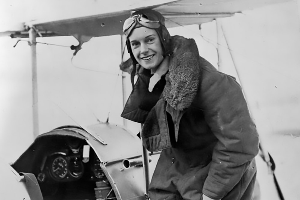 Ч/б фотографія Джин Баттен у літальному одязі поруч із її літаком