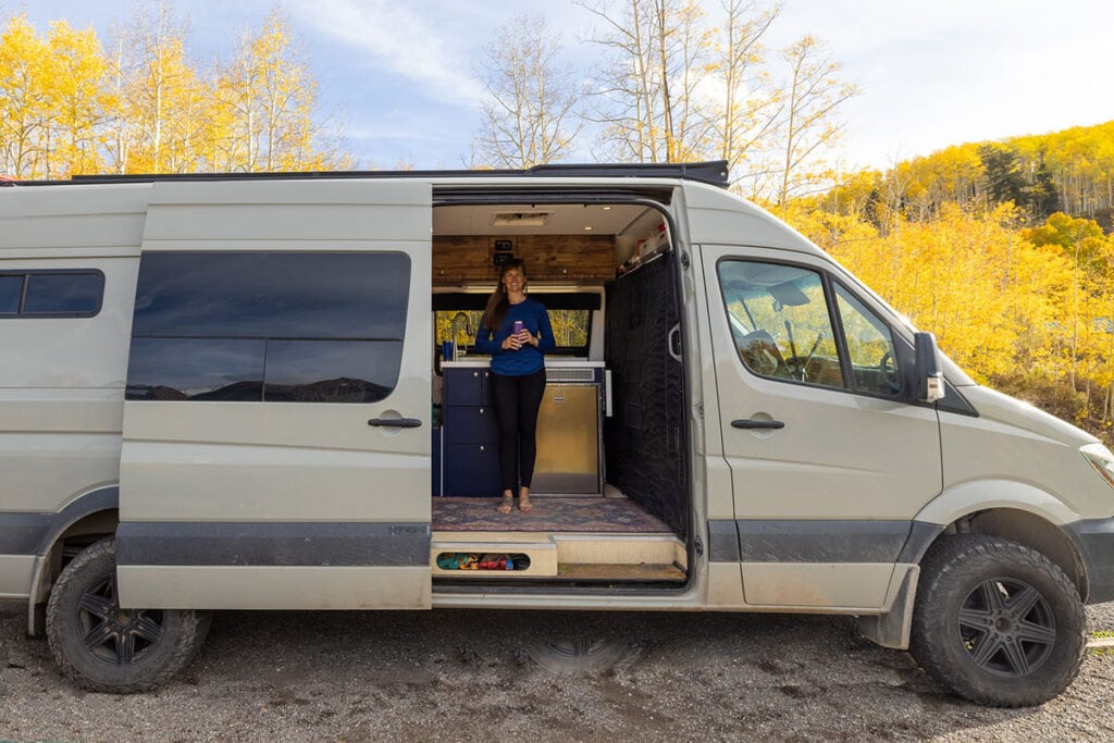  Засновник Bearfoot Theory Крістен Бор стоїть у дверях кемпер Sprinter у Теллурайді, штат Колорадо, із золотистими осиками позаду фургона